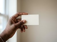 Male hand with dark skin tone holding blank white business card or paper against neutral beige wall with natural window light, suitable for branding, corporate identity, and marketing mockups.