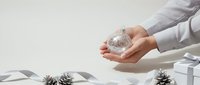 Hands holding crystal snow globe with winter scene, surrounded by pine cones and gift boxes on white surface for holiday decoration and seasonal celebration.