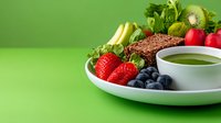 Fresh healthy breakfast with berries, kiwi, banana, matcha green tea, and whole grain bread on white plate against vibrant green background.