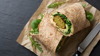 Whole wheat wrap sandwich filled with green vegetables and hummus, sliced in half on kraft paper with fresh greens, ready for lunch or meal prep photography.