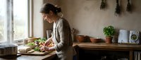 Young woman preparing fresh salad in rustic kitchen with herbs hanging on wall. Healthy cooking lifestyle concept for organic food preparation.