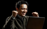 Asian man celebrating success with laptop, raising fists in victory gesture against black background, expressing joy and achievement.