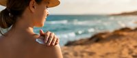 Woman applying sunscreen lotion on shoulder at beach during sunny day, protecting skin from UV rays and sun damage for summer vacation skincare routine.