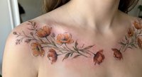 Delicate floral tattoo across woman's chest and shoulders featuring orange wildflowers and botanical elements on fair skin in natural lighting.