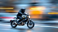 Male motorcyclist in black leather riding sports bike on urban street with motion blur effect creating sense of speed and dynamic movement.
