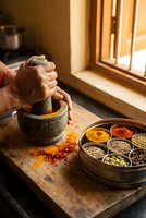 Hands grinding spices with traditional stone mortar and pestle in rustic kitchen with colorful spice collection in wooden tray for authentic cooking.
