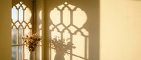 Warm sunlight streaming through ornate gothic windows casting decorative shadows on cream wall with dried flowers creating peaceful interior atmosphere.