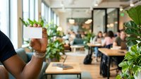 Employee holding blank white business card in modern office workspace with green plants and colleagues working at desks in background.