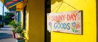 Colorful tropical storefront with handmade wooden sign reading Sunny Day Goods, featuring bright yellow walls and vibrant street scene for retail business concepts.