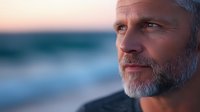 Confident mature Caucasian man with gray beard gazing thoughtfully at sunset on beach, profile view against blurred ocean background shows wisdom and contemplation.