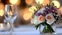 Elegant wedding table setting with crystal wineglass and fresh floral centerpiece featuring peach roses and purple flowers against soft bokeh lights background.
