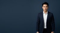 Young Asian male professional in dark navy blazer and white dress shirt standing against blue background, suitable for corporate, business, and corporate portrait use.
