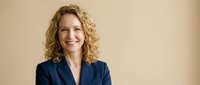 Professional middle-aged Caucasian businesswoman with curly blonde hair smiling confidently in navy blue blazer against neutral beige background.