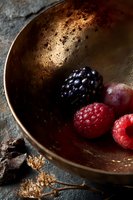 Fresh raspberries and blackberry in rustic bronze bowl on dark stone background with chocolate pieces and dried flowers, moody food photography styling.