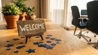 Wooden welcome sign on office desk with blue star confetti and potted plants creating warm professional workspace atmosphere for business greeting.