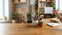Creative professional workspace with wooden desk, holding blank white card, paintbrushes in ceramic holder, potted plants, and shelving with books and decor in bright studio.