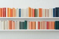 Minimalist bookshelf with colorful book spines arranged in pastel and muted tones on white shelves against light wall, creating modern home library aesthetic.