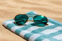 Stylish aviator sunglasses with dark green lenses resting on striped beach towel against golden sand background for summer vacation and travel concepts.