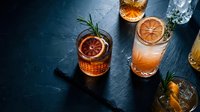 Whiskey cocktails with orange garnish and rosemary on dark slate background. Premium spirits and craft mixology for bar menu design and beverage photography.