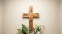 Wooden cross with natural grain pattern displayed on neutral wall with green fern leaves for religious decoration and spiritual worship concepts.