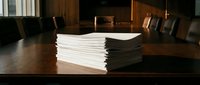 Large stack of white paper documents on wooden conference table in professional boardroom setting for business meetings and corporate presentations.