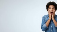 Young man with dark curly hair and medium skin tone wearing denim shirt, expressing shock and surprise with hands on face against white background for advertising and marketing campaigns.