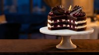 Decadent chocolate layer cake with rich frosting and berry filling displayed on elegant white pedestal stand in warm bakery setting.