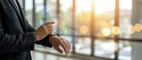 Businessman in dark suit holding transparent digital tablet with glowing data visualization and financial charts, modern office with blurred city skyline background at sunset, technology and business analytics concept.