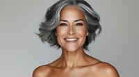 Confident mature Hispanic woman with silver gray hair and natural makeup smiling warmly against neutral background, showcasing beauty in aging gracefully.