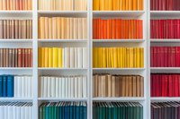 Colorful books arranged by spectrum on white shelves create modern rainbow pattern. Books spines in blue, yellow, orange, and red tones form organized library display.