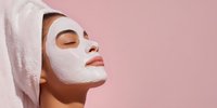 Young woman with white facial mask and towel on head relaxing with eyes closed against pink background for skincare and beauty routine.