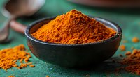 Vibrant orange turmeric powder in black ceramic bowl on green rustic background with scattered spice. Fresh ground aromatic seasoning for cooking and wellness.