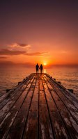 Couple standing on wooden pier at sunset over ocean, silhouetted against golden sky with dramatic clouds and calm water, romantic travel destination for relationships and vacations.