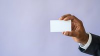 Close-up of male hand with dark skin tone wearing business suit holding blank white business card against purple background for corporate branding.