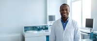 Smiling African American male doctor in white lab coat standing confidently in modern medical clinic with equipment and computers in background.