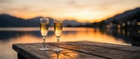 Two champagne glasses on wooden dock at sunset with mountain lake reflection creating romantic evening atmosphere for celebration and luxury dining.