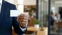 Professional man in dark suit holding blank white business card in modern office environment with blurred colleagues in background, suitable for corporate branding and business networking.