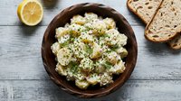 Creamy potato salad with fresh herbs and lemon in rustic bowl, served with toasted bread slices on wooden table for lunch or dinner meal preparation.