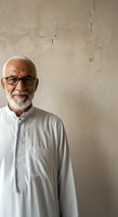 Senior man with white hair and beard wearing eyeglasses and light gray traditional tunic shirt, standing against neutral textured wall background for portrait use.