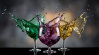 Colorful liquid splashing from three wine glasses in green, purple and yellow creating dynamic motion against dark background.
