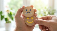 Hands holding wooden emotion faces showing sad and happy expressions for mental health awareness and emotional wellbeing concepts in bright indoor setting.