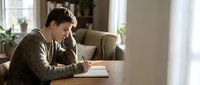 Young Caucasian man in sweater writing in notebook at wooden table in bright modern living room with natural light streaming through windows.