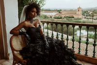 Elegant African American bride in black feathered gown sitting on Mediterranean balcony overlooking scenic countryside for luxury wedding photography.
