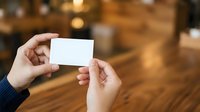 Two hands exchanging blank white business card against warm wooden interior background with bokeh lighting, suitable for corporate branding and networking concepts.