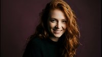 Young redhead woman with curly hair smiling warmly in dark sweater against moody background for lifestyle and portrait photography projects.