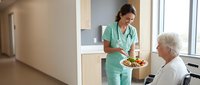 Young female healthcare worker in green scrubs serving healthy meal to elderly Caucasian patient in modern hospital room with natural lighting.