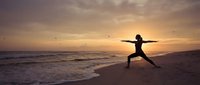 Woman practicing warrior yoga pose on beach at sunset with golden sky and ocean waves creating peaceful meditation scene.