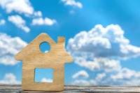 Wooden house silhouette against blue sky with white fluffy clouds. Simple minimalist home symbol on rustic wooden surface represents real estate and property concepts.