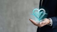 Young adult male with light skin tone wearing dark business attire holding glowing turquoise heart symbol in open palm, representing love, care, charity and digital connection for corporate and healthcare applications.