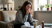 Young Caucasian woman with flu symptoms blowing nose with tissue while sitting on couch at home surrounded by medicine bottles and tea cup.